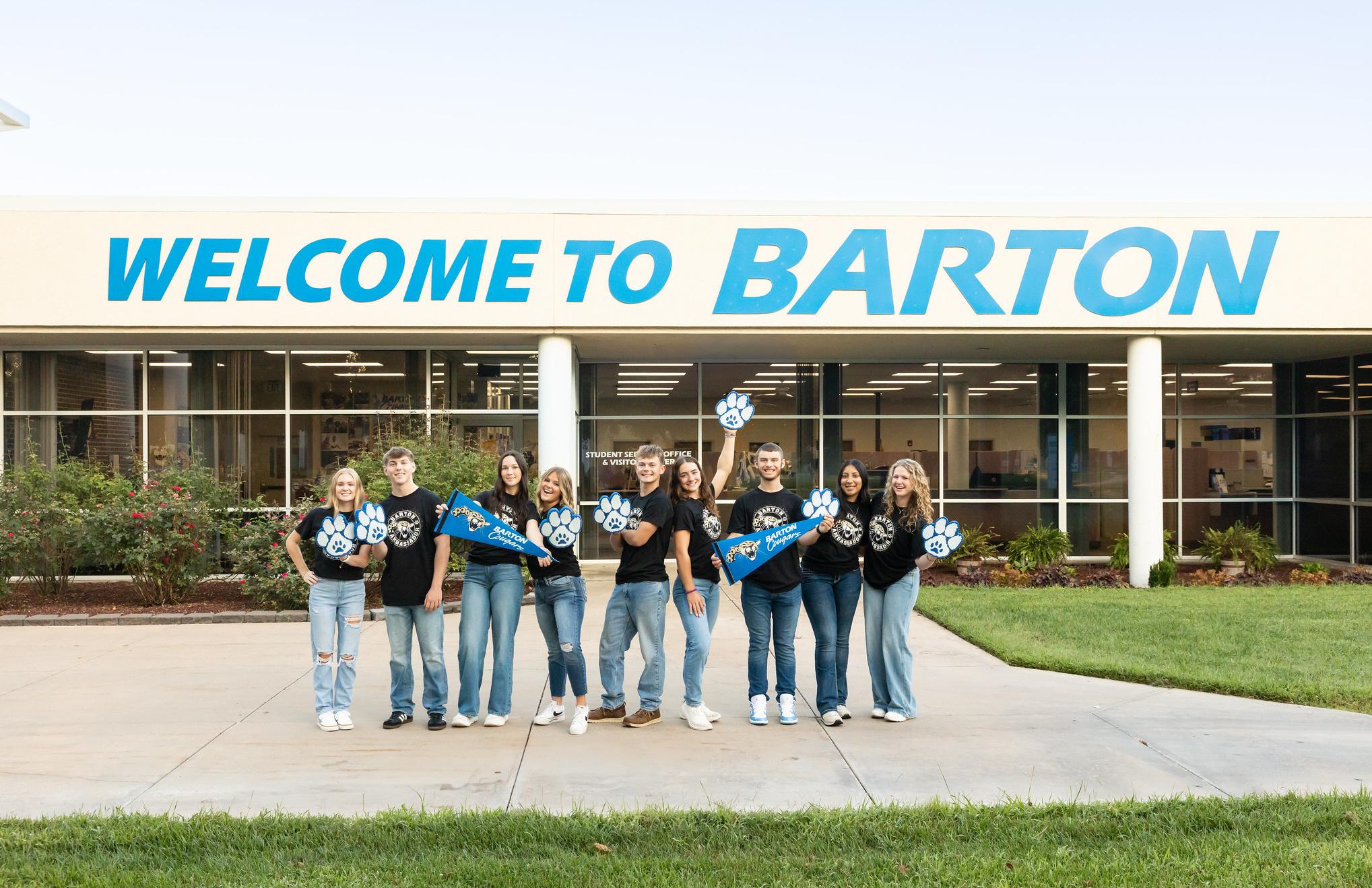 2025 Ambassadors smile in front of the Welcome to Barton signage on campus
